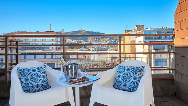 A balcony with two white chairs, a small table, a bottle of champagne, and a plate of snacks. - Mercure Barcelona Condor (Barcelona)