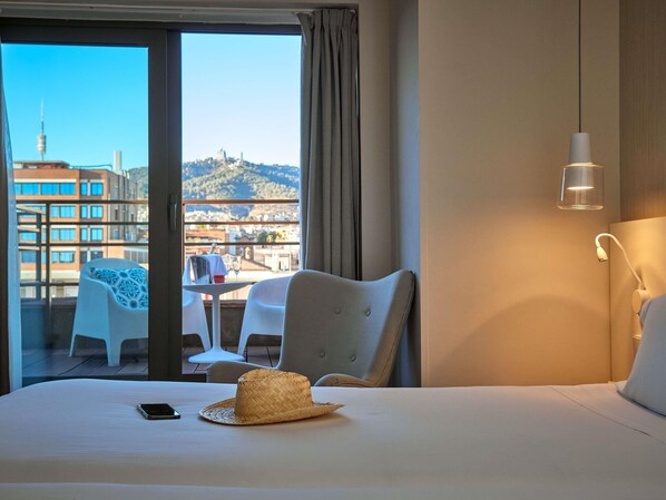 Una habitación de hotel con cama, una mesa de comedor con sillas, un balcón con vistas a edificios y una colina, y una lámpara colgante.
