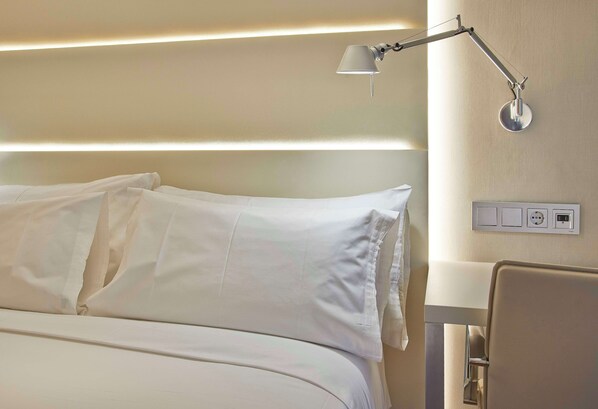 Una cama bien hecha con sábanas blancas, una mesita de noche y una lámpara fijada a la pared.