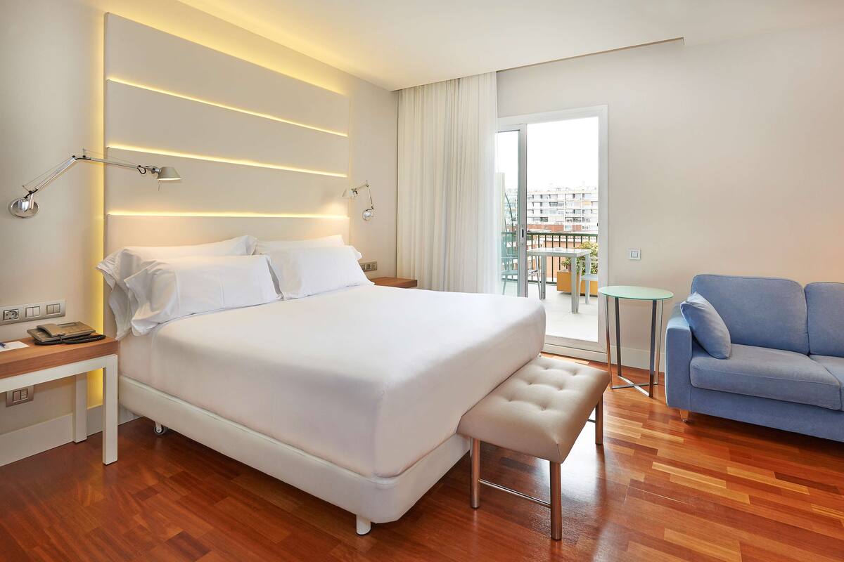 Una habitación de hotel moderna con una cama grande, una mesita pequeña, un sofá azul y un balcón con vistas a la ciudad.