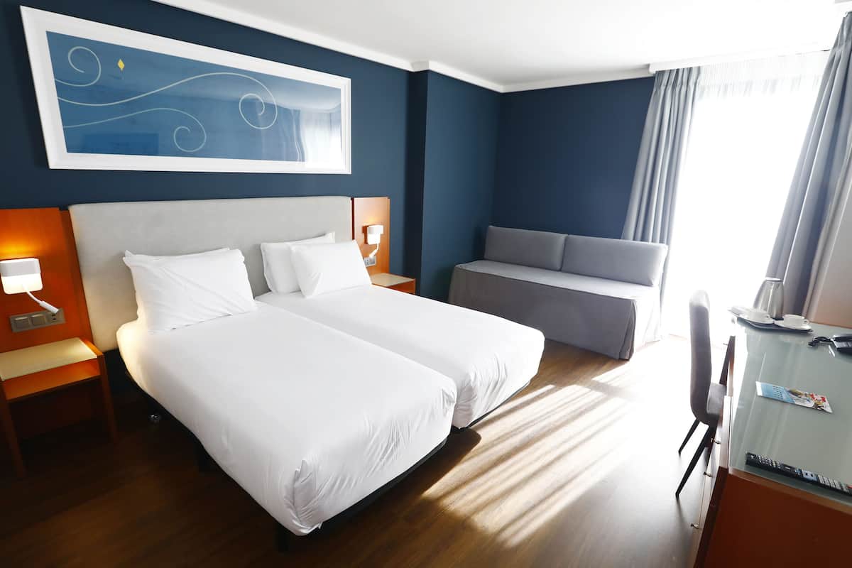 A hotel room with two beds, a sofa, a desk with a phone and remote controls, and a painting on the wall.