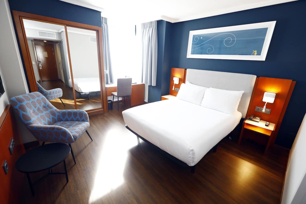 A hotel room with a large bed, a desk, a chair, and a mirror.