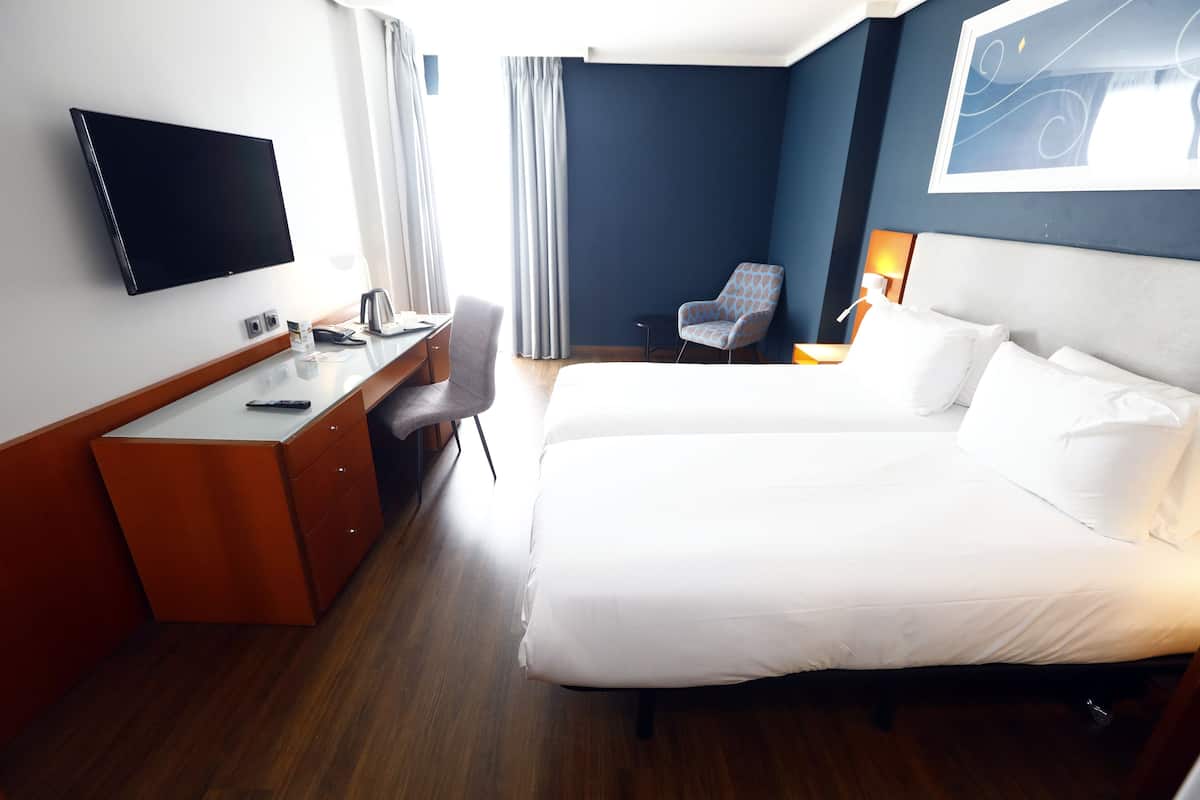 A hotel room with a bed, a desk with a chair, a TV, and a window with curtains.