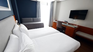 A hotel room with two beds, a desk, a chair, a TV, and a window with curtains.