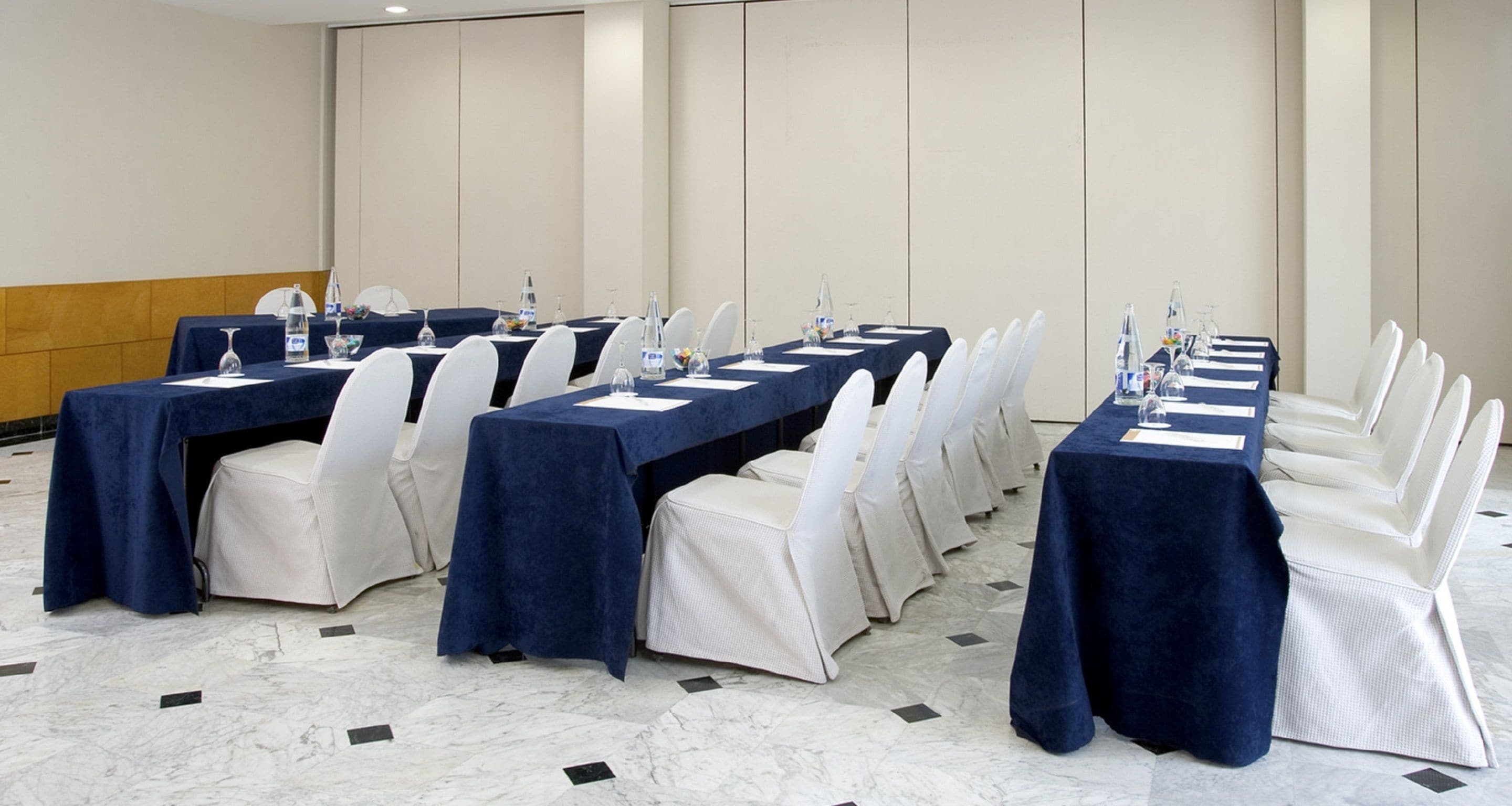 Un salón de conferencias con largas mesas, sillas blancas y manteles azules.