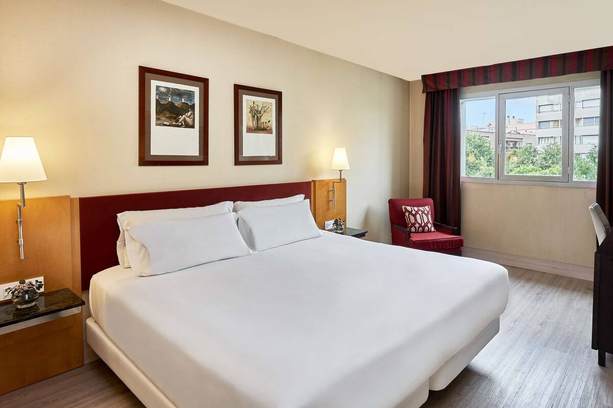 A hotel room with a large bed, two wall-mounted lamps, a red chair, a small table with a bottle and glasses, and two framed pictures on the wall.