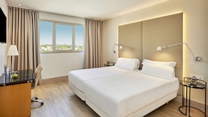 A hotel room with a large bed, two bedside lamps, a desk with a lamp, a chair, and a view of the city through the window. - NH Ciutat de Reus (Reus)