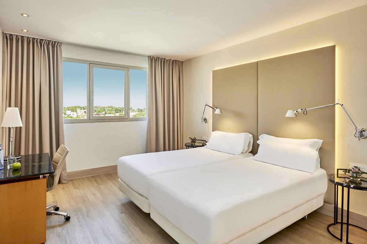 A hotel room with a large bed, two bedside lamps, a desk with a lamp, a chair, and a view of the city through the window.