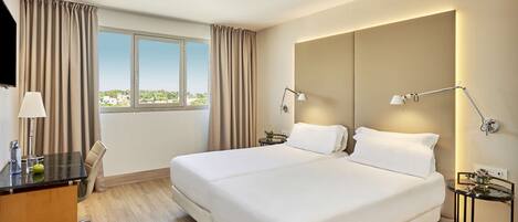 Quarto de hotel com uma cama grande, duas luminárias de cabeceira, uma escrivaninha com abajur, uma cadeira e vista para a cidade pela janela.