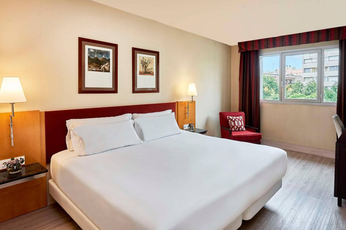 A hotel room with a large bed, two framed artworks on the wall, a red armchair, a desk with a lamp, and a window with curtains.
