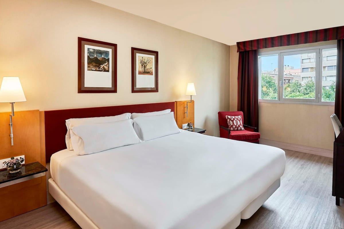 A hotel room with a large bed, two framed artworks on the wall, a red armchair, a desk with a lamp, and a window with curtains.