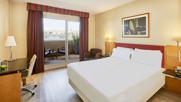 Quarto de hotel com uma cama grande, uma escrivaninha com cadeira, uma mesinha, duas luminárias e uma janela com cortinas.