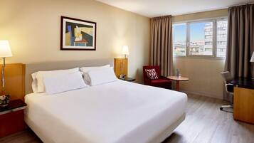 Una habitación de hotel con una cama grande, dos lámparas de noche, una mesita con una silla, un escritorio y una ventana con cortinas.