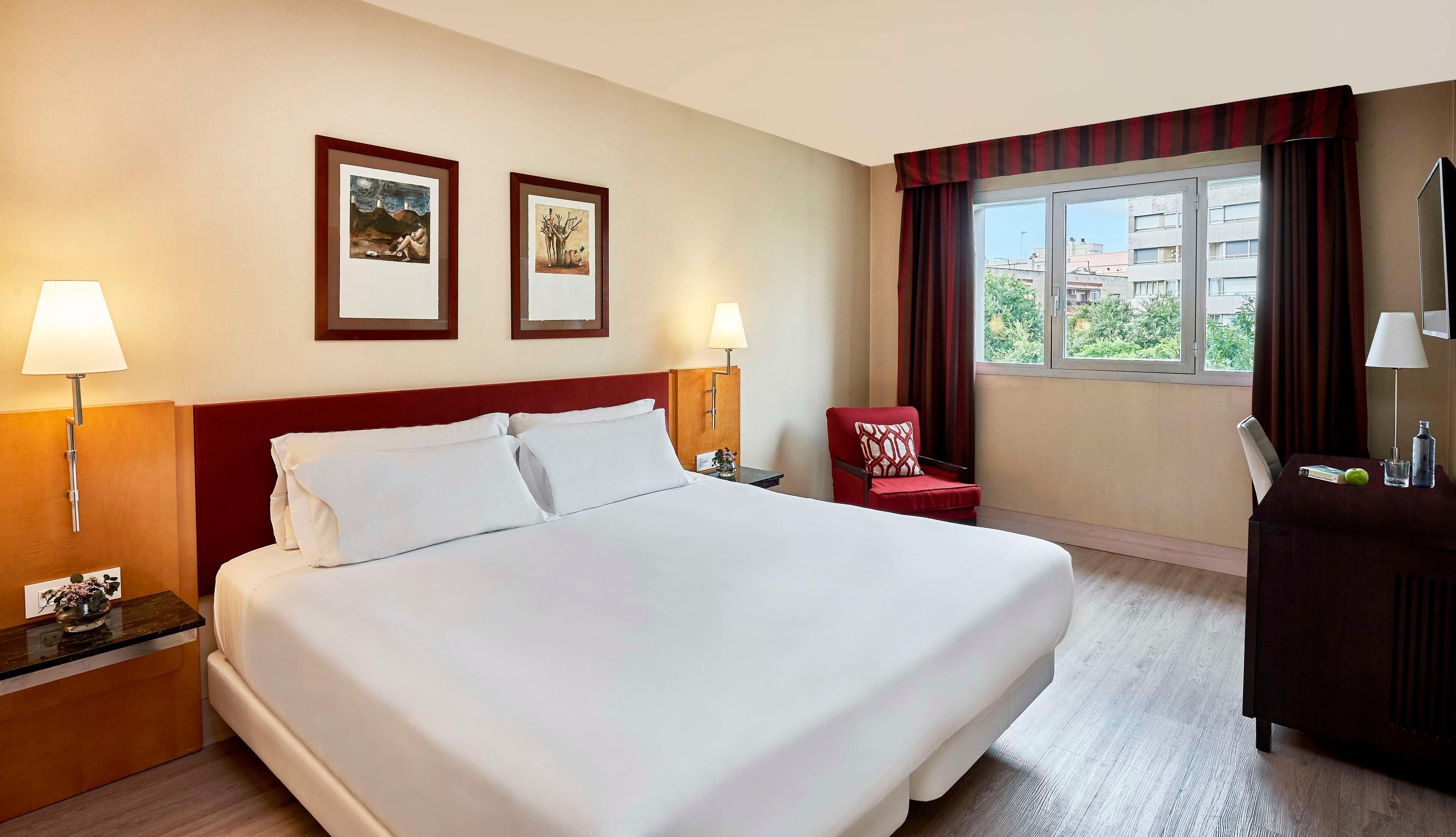 A hotel room with a large bed, two framed artworks on the wall, a red armchair, a desk with a lamp, and a window with curtains.