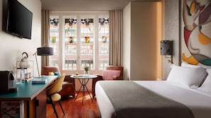 Una habitación de hotel con una cama, un escritorio con lámpara, una silla, una mesita con un florero y un televisor montado en la pared.
