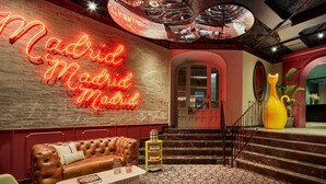A cozy interior with a neon sign reading "Madrid Madrid," a leather sofa, a coffee table, and a staircase leading to an upper level.
