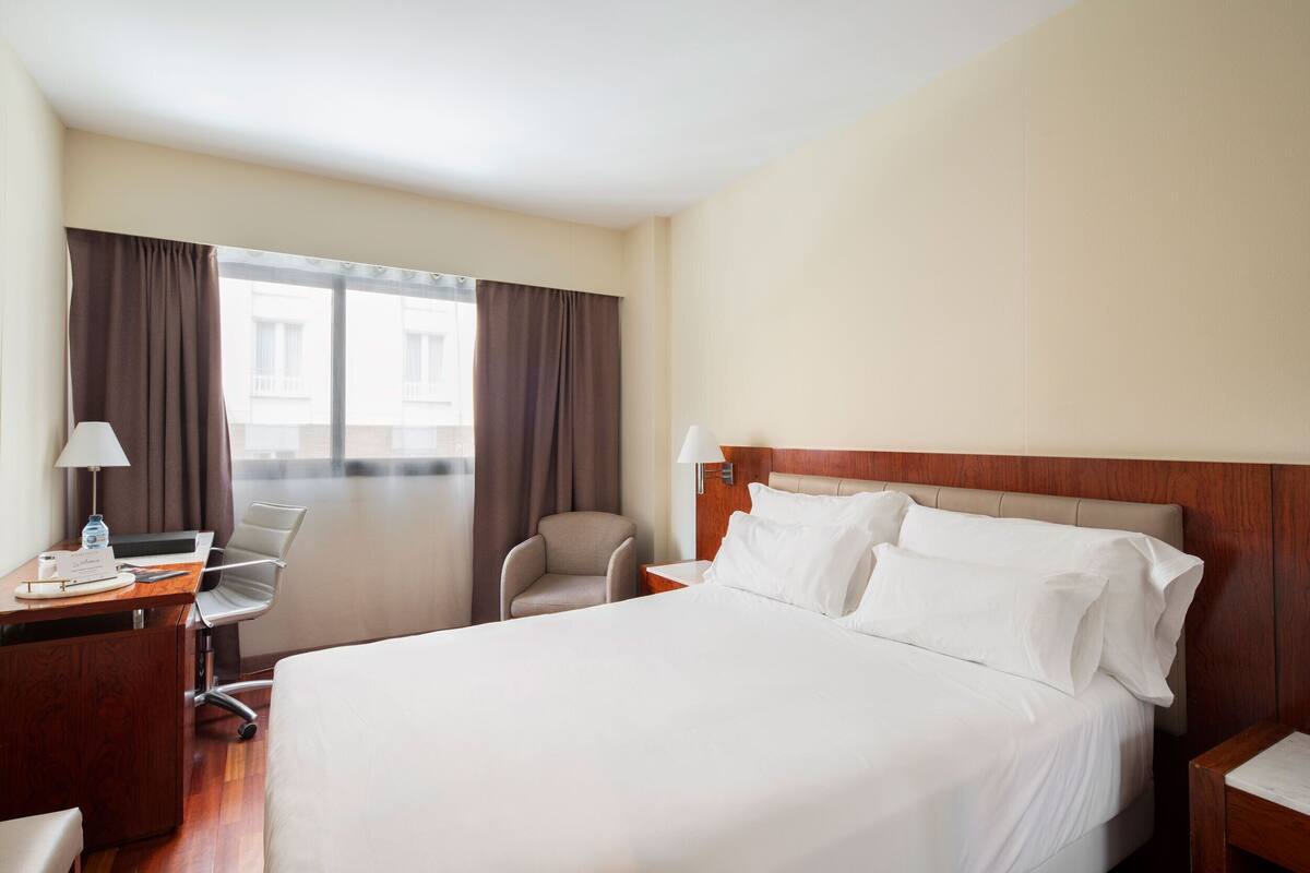 A hotel room with a bed, desk, chair, lamp, and large window with curtains.