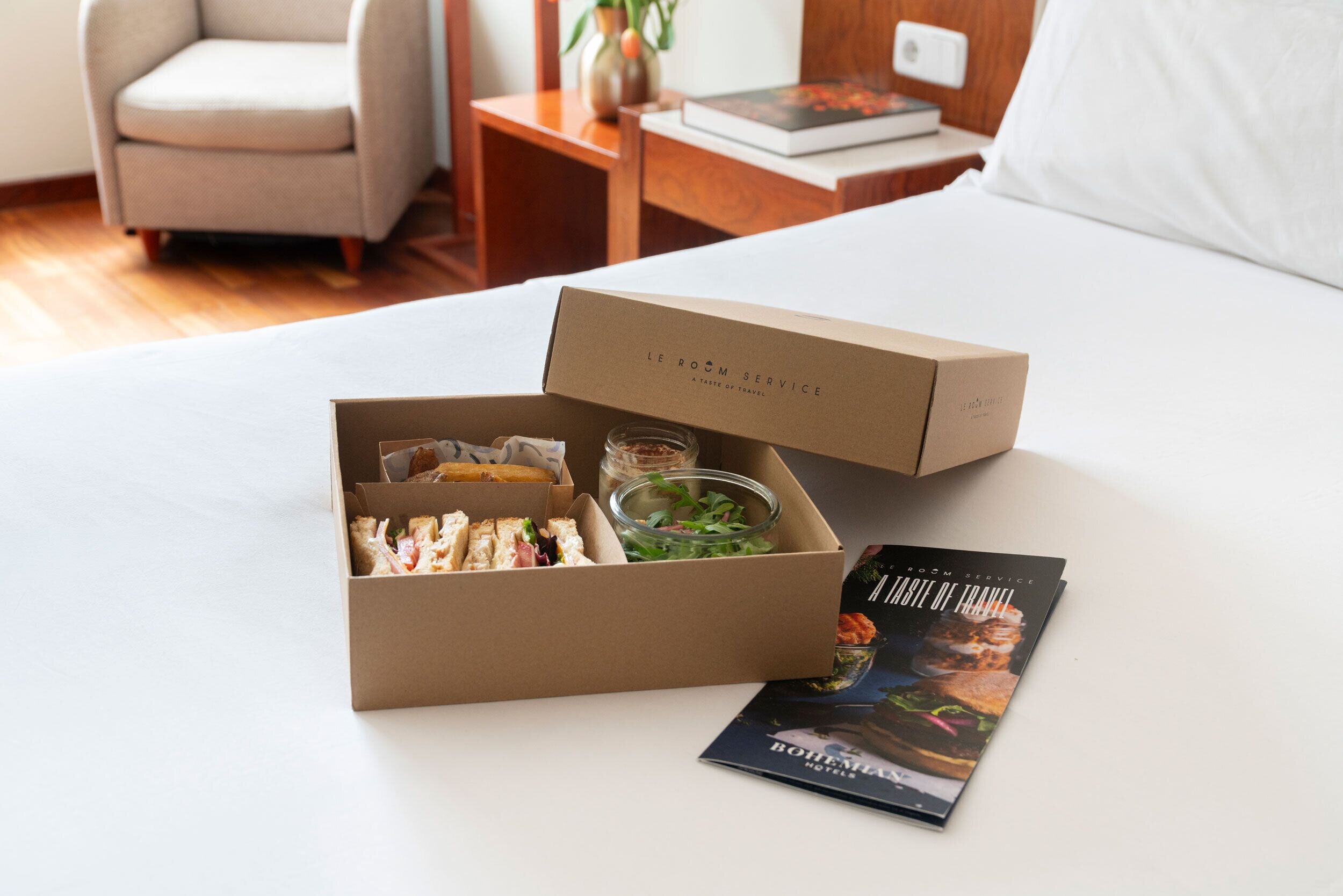 Een keurig opgemaakt ontbijtboxje met belegde broodjes, een salade en een drankje op een bed, met naast het bed een boek met de titel 'A Taste of Travel'.