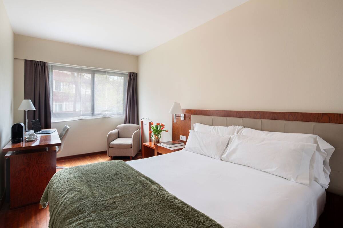 A hotel room with a bed, desk, chair, lamp, and window.