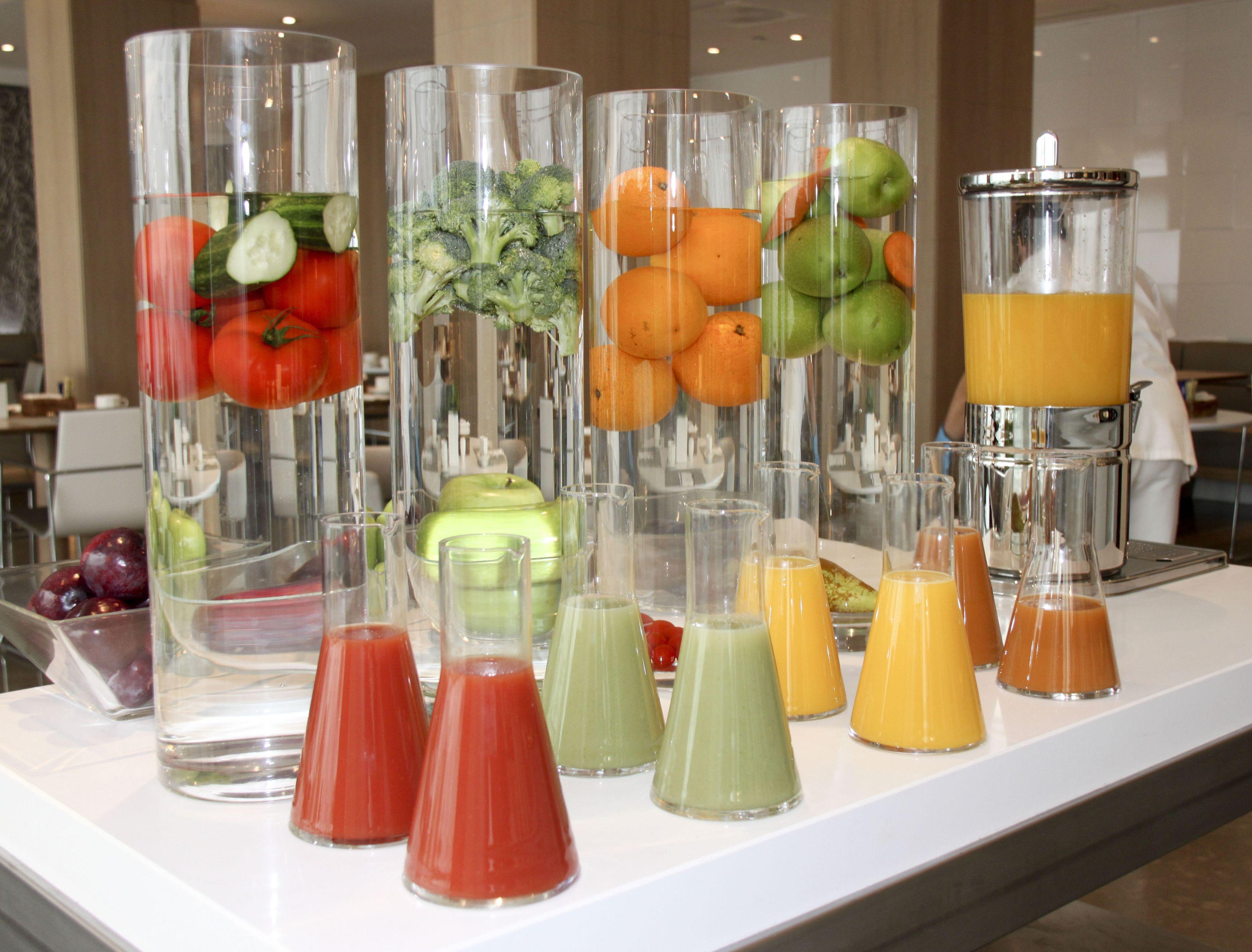 Un bufé con diversas frutas y jugos de vegetales en vasos transparentes.