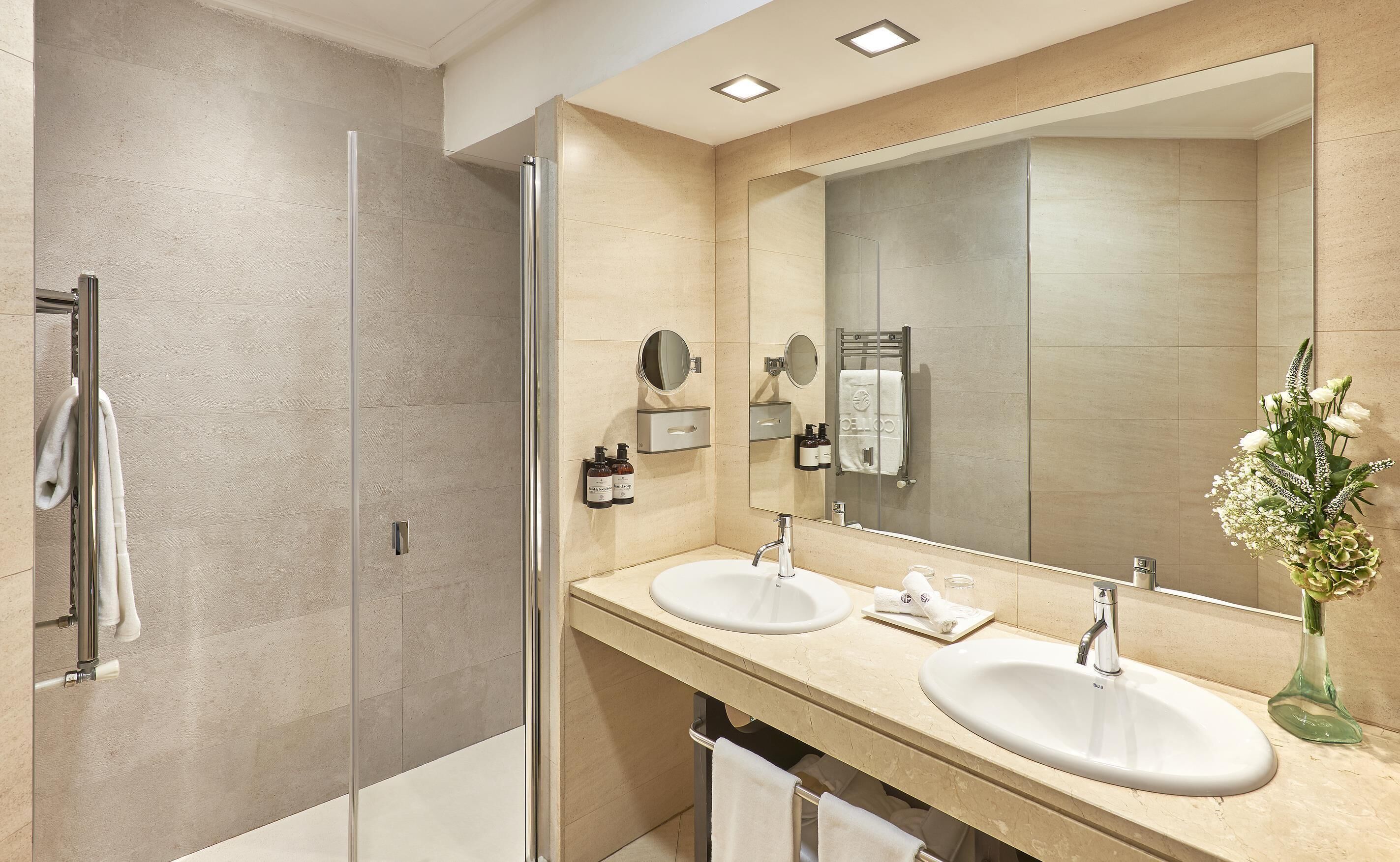 A modern bathroom with a glass shower enclosure, two sinks, a large mirror, and a vase with flowers.