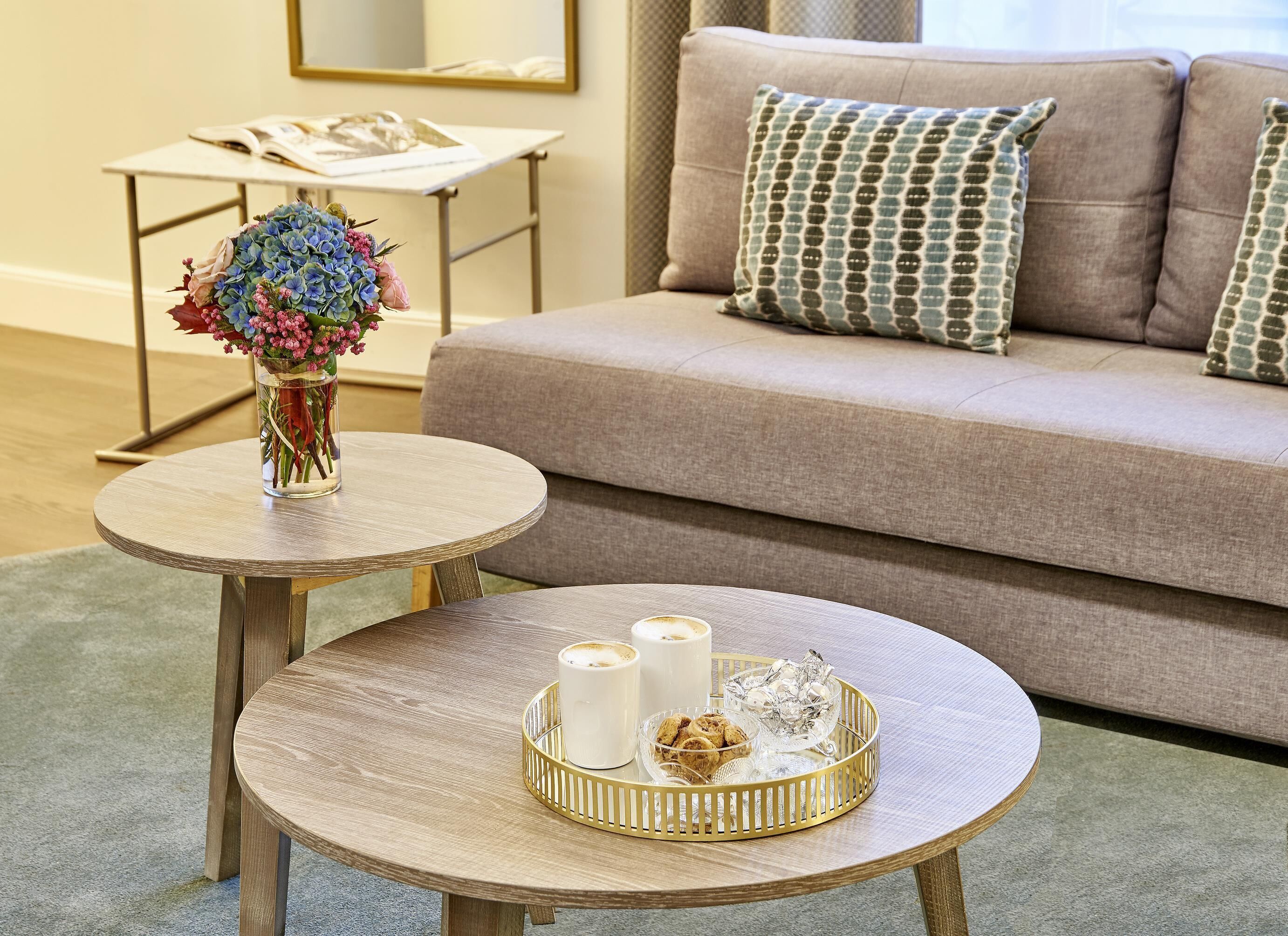 Un salon avec un canapé beige, deux petites tables rondes en bois, dont l’une est ornée d’un vase de fleurs et l’autre de bougies et d’un plateau de biscuits.
