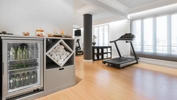 A gym area with a treadmill, a refrigerator stocked with beverages, and a window with a view of buildings.