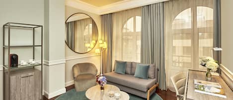Una habitación de hotel moderna con sofá, dos sillones, una mesita redonda, un mueble bajo y una mesa de comedor con un jarrón de flores.