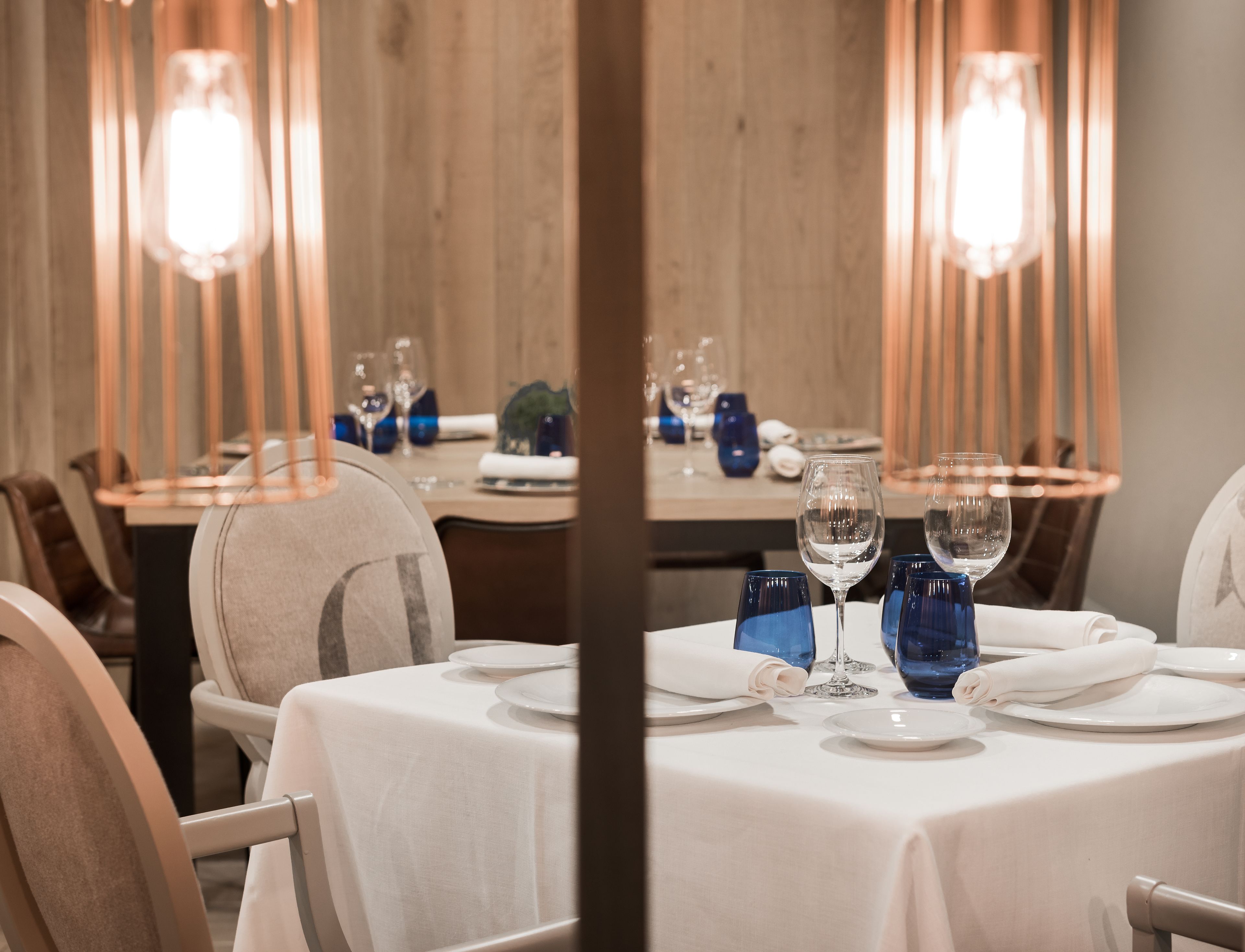 Comedor con mesas dispuestas para una comida, con cristalería azul y manteles blancos.