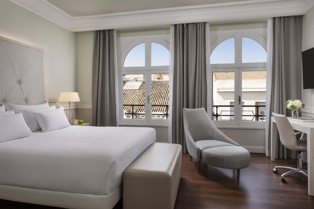 Habitación de hotel con una cama grande, un escritorio, una silla y vistas de la ciudad a través de ventanales arqueados.