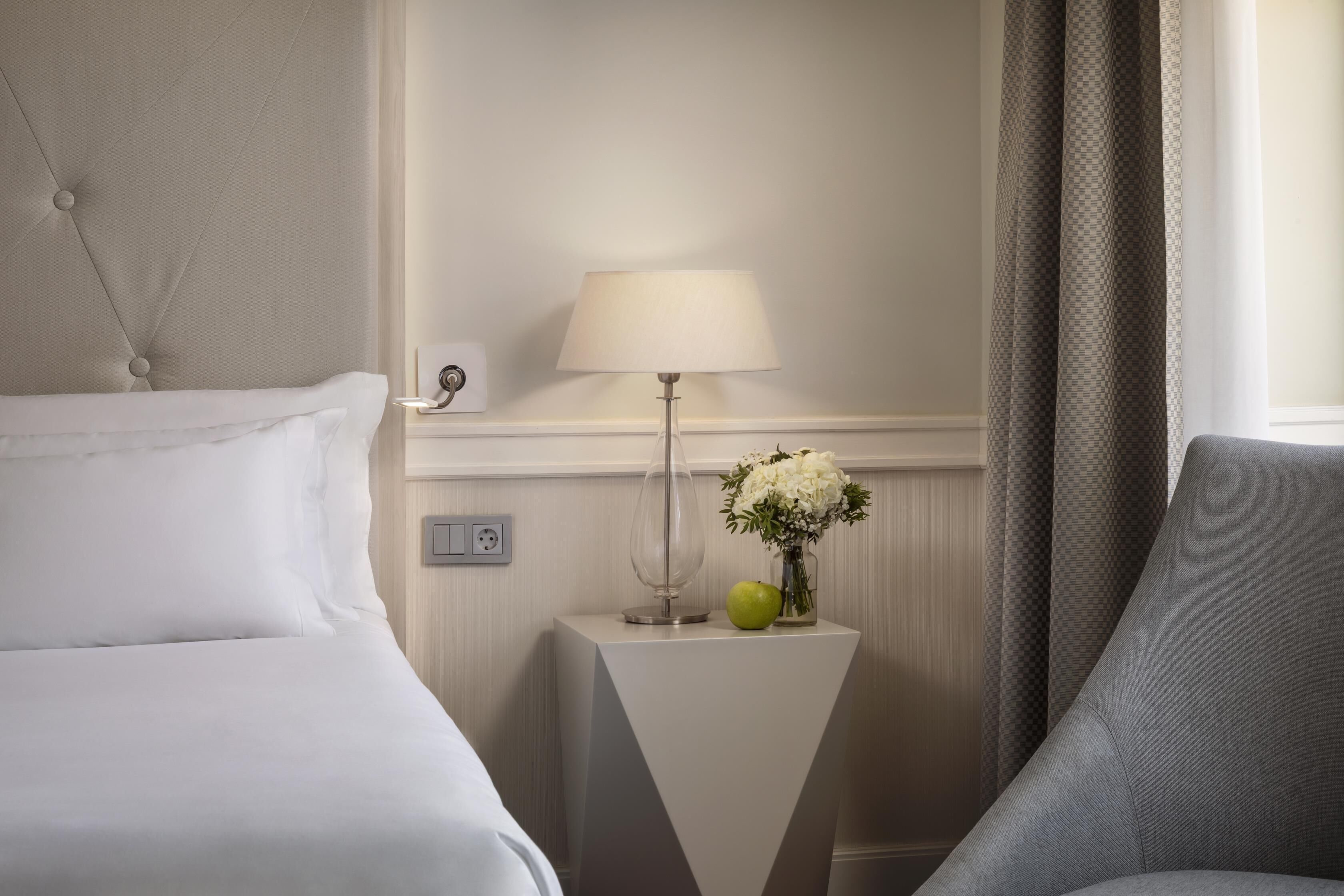 A hotel room with a bed, a nightstand with a lamp, a vase with flowers, and a chair.
