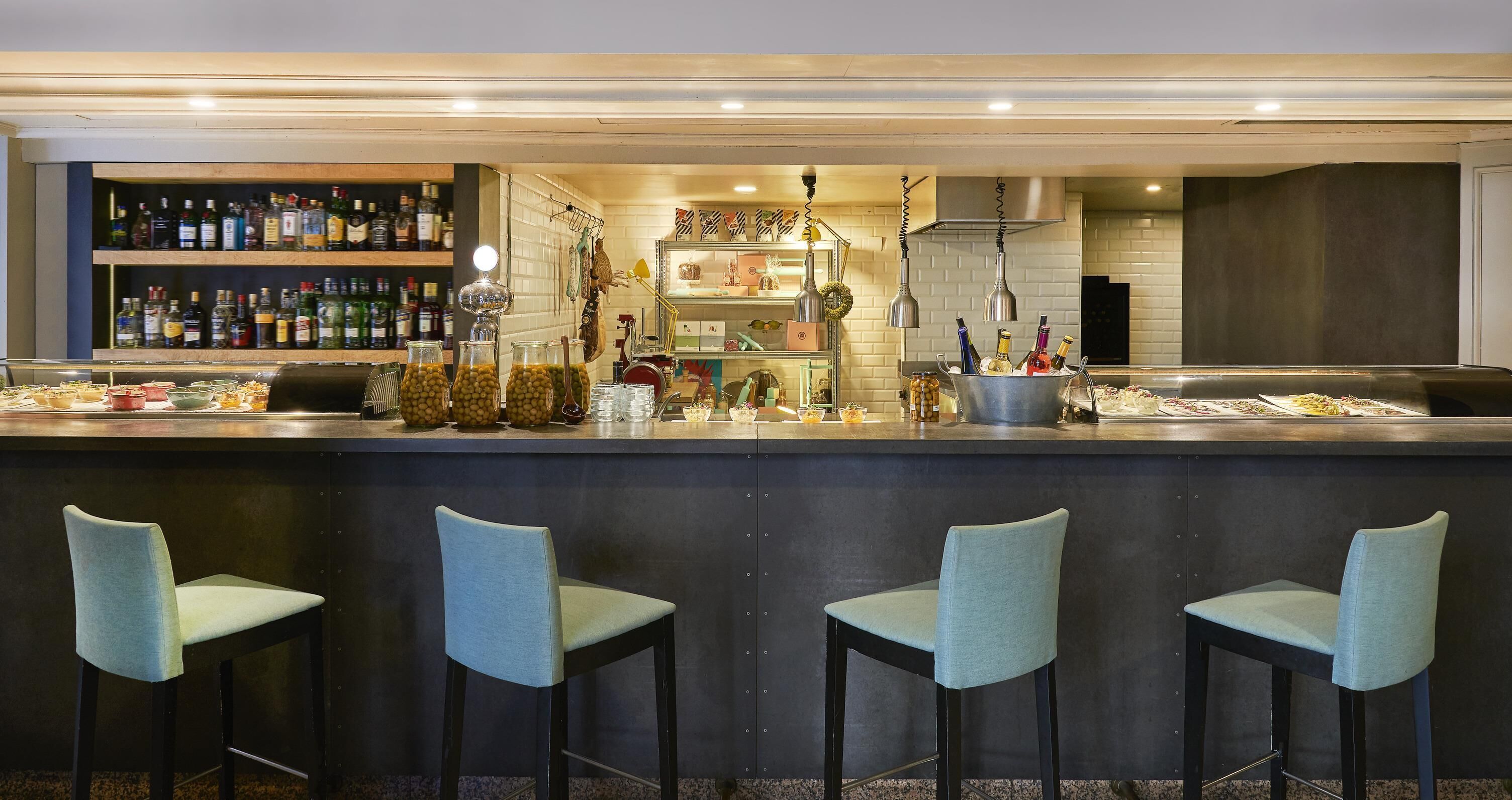 Zona de bar con mostrador, estantes llenos de botellas de licores y tres taburetes.
