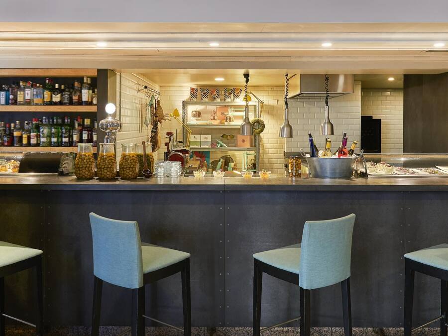 Zona de bar con mostrador, estantes llenos de botellas de licores y tres taburetes.