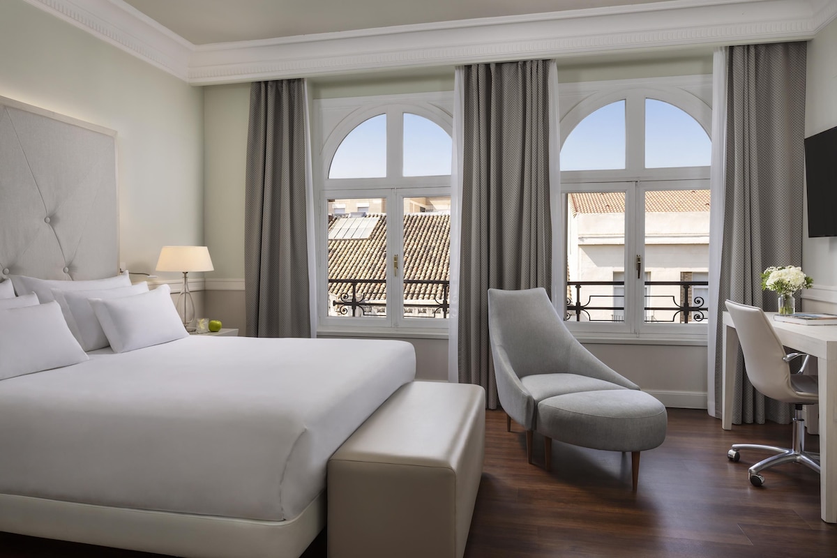 A hotel room with a large bed, a desk, and a chair. There is a window with curtains and a view of buildings outside.