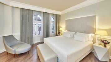 Un cuarto de hotel con una cama grande, una silla, una mesita de noche con lámpara y vistas al exterior a través de una ventana con cortinas.