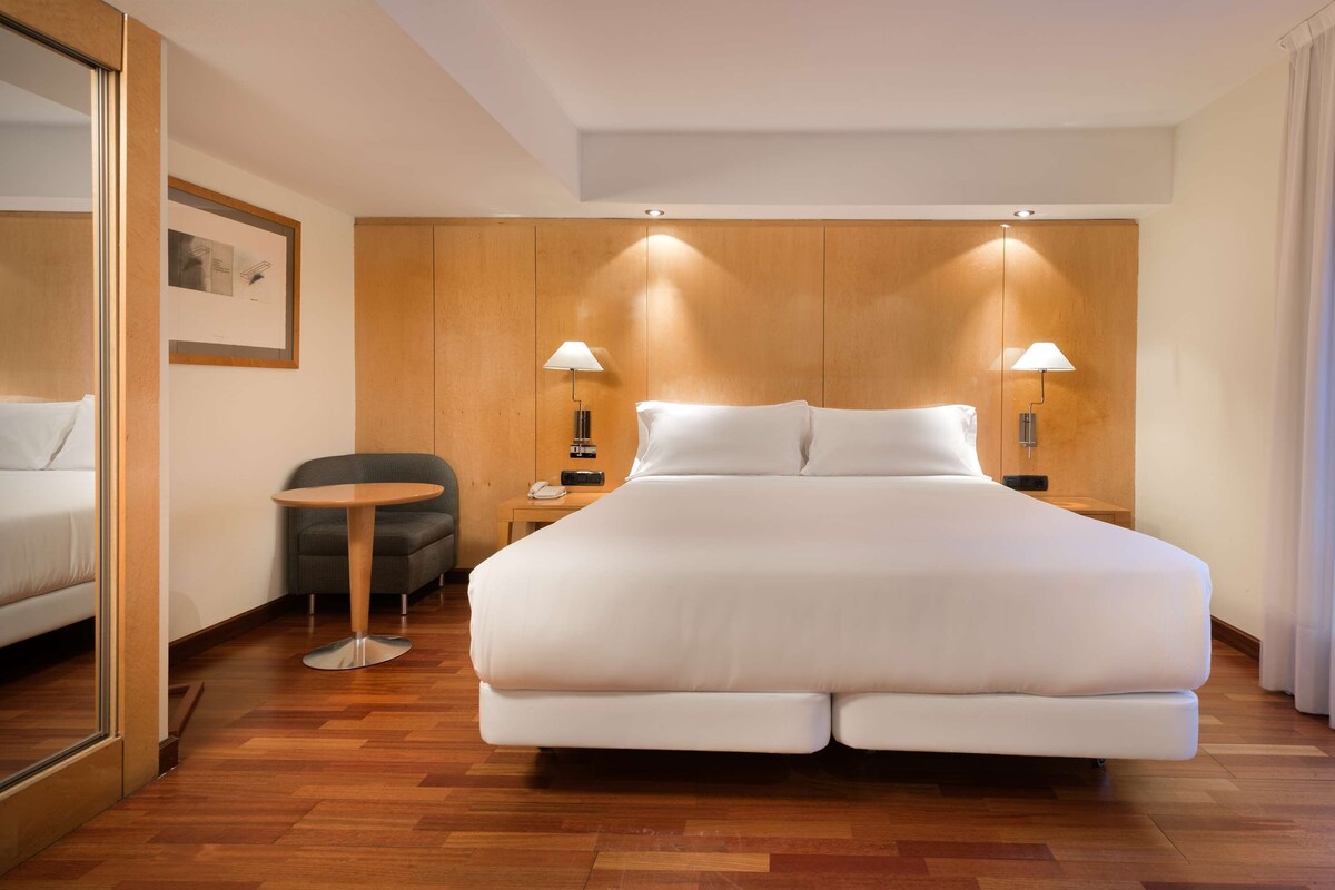 A hotel room with a large bed, a small round table, a chair, a desk with a lamp, and a telephone.