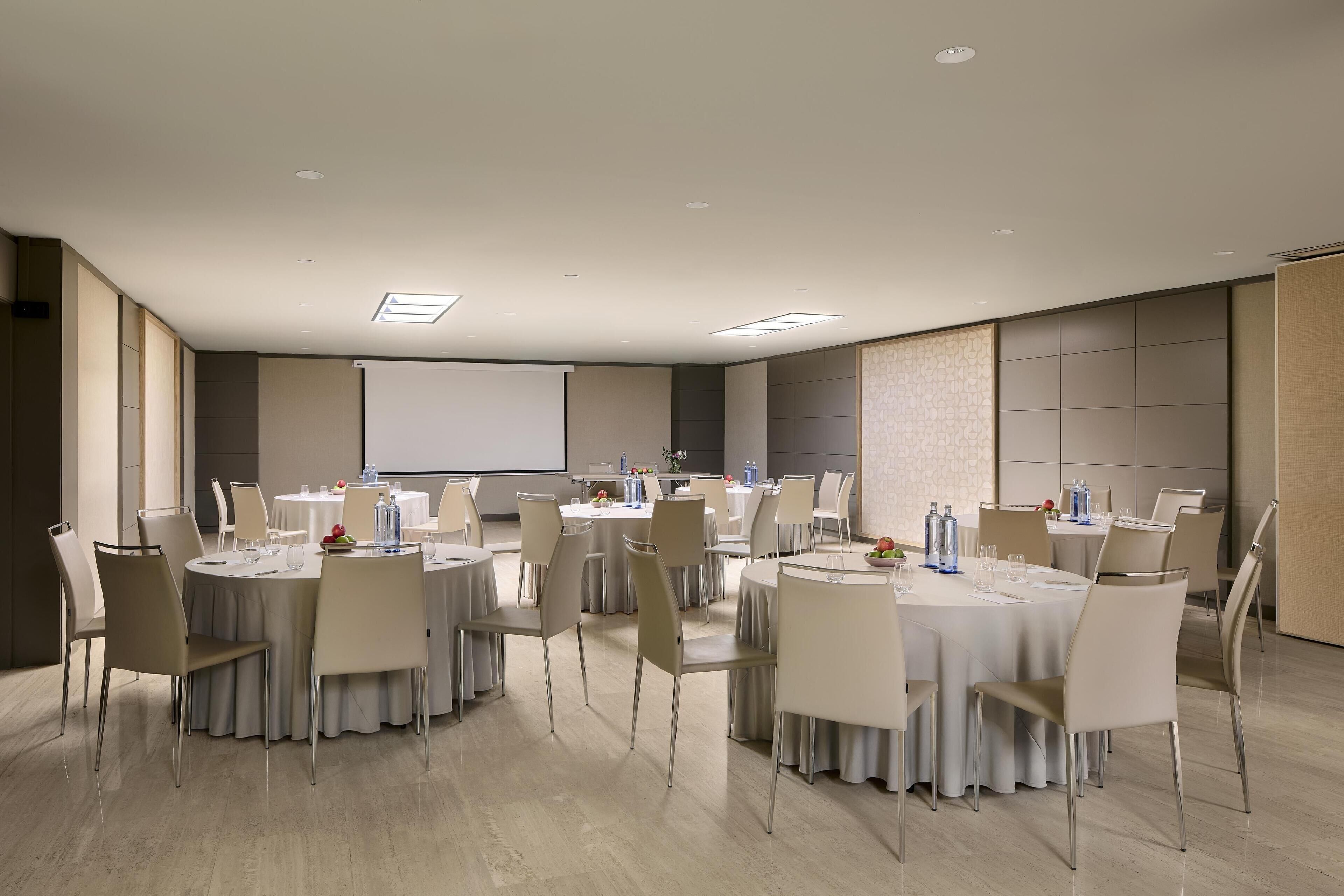Un salón de conferencias con mesas redondas, sillas y una pantalla de proyector.