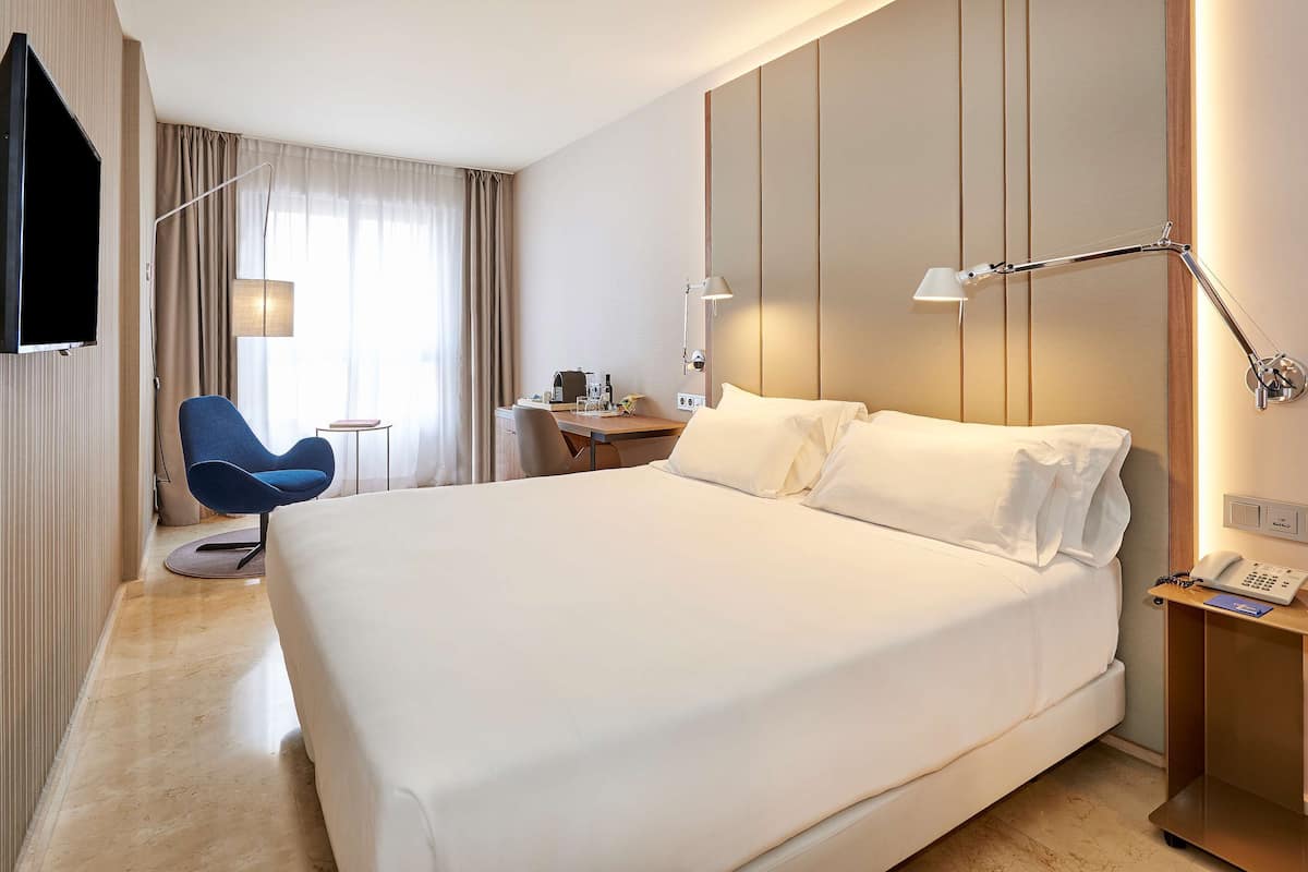 A hotel room with a large bed, a desk, a chair, a TV, and a bedside table with a lamp.
