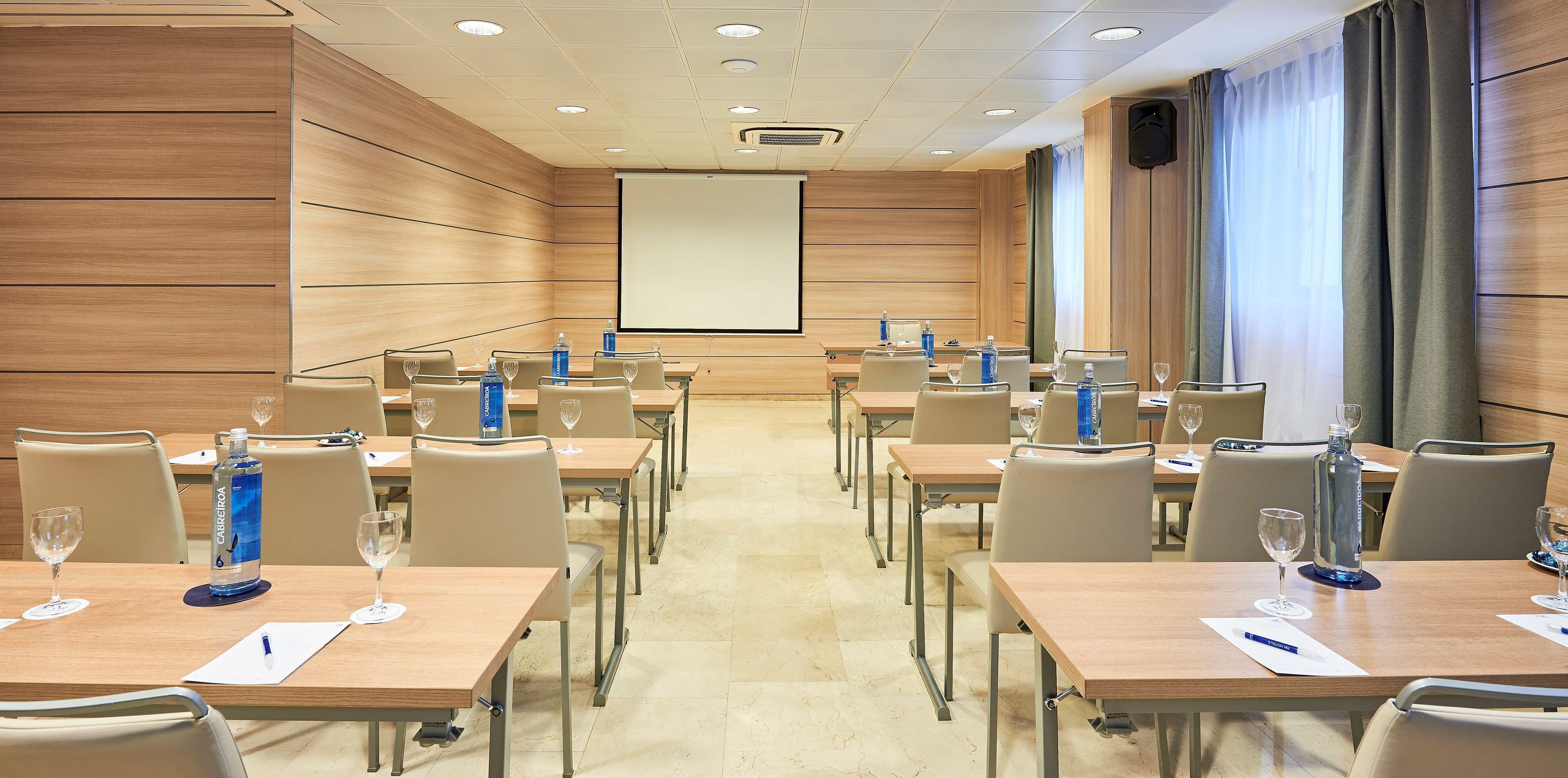 Una sala de conferencias con paneles de madera, una pantalla de proyección grande y varias mesas con sillas y botellas de agua.