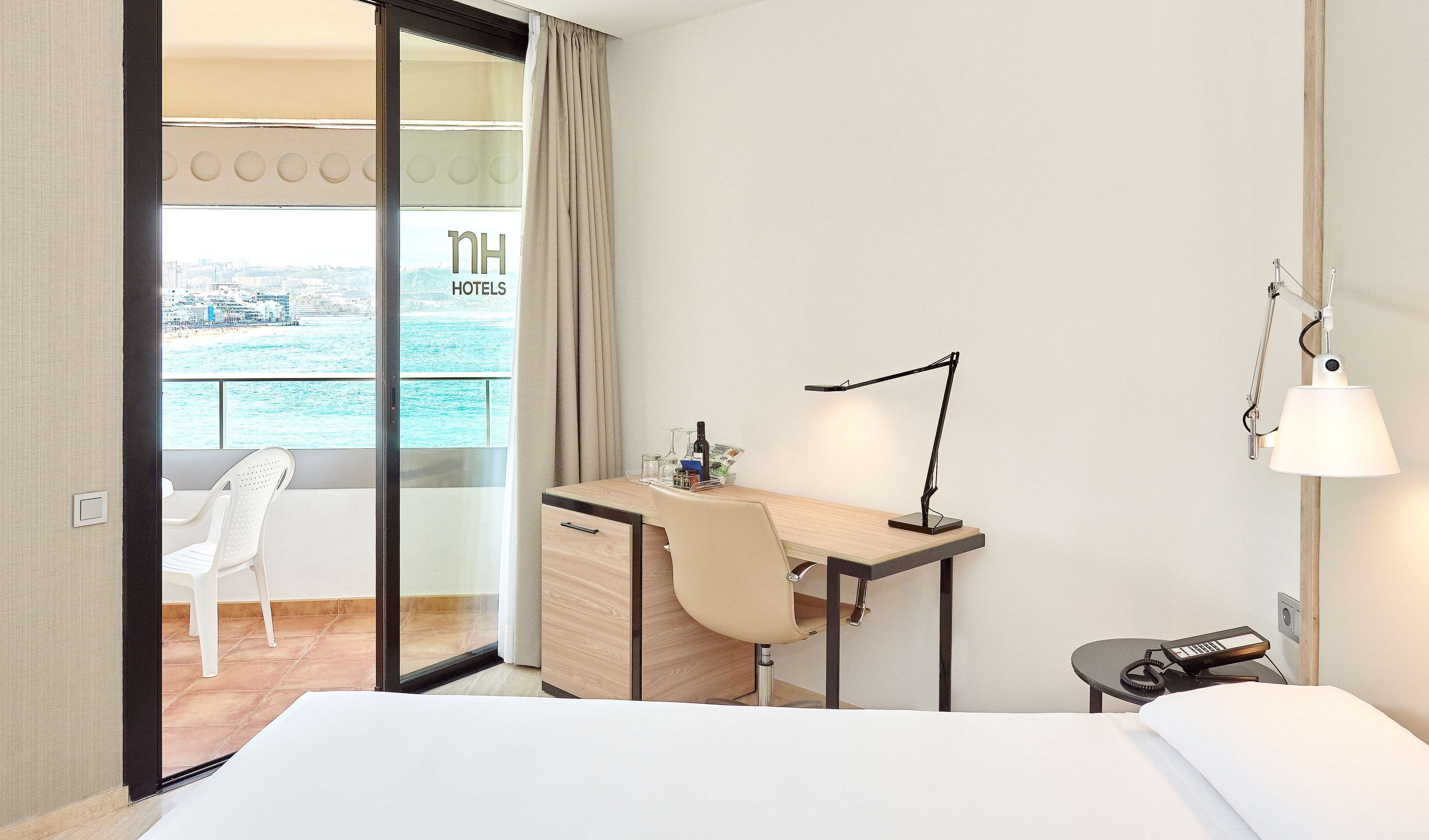 Ein Hotelzimmer mit Bett, Schreibtisch mit Lampe, Stuhl, kleinem Tisch mit Telefon und Meerblick durch eine Glastür.
