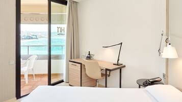 A hotel room with a bed, a desk with a lamp, a chair, a small table with a phone, and a view of the sea through a glass door.