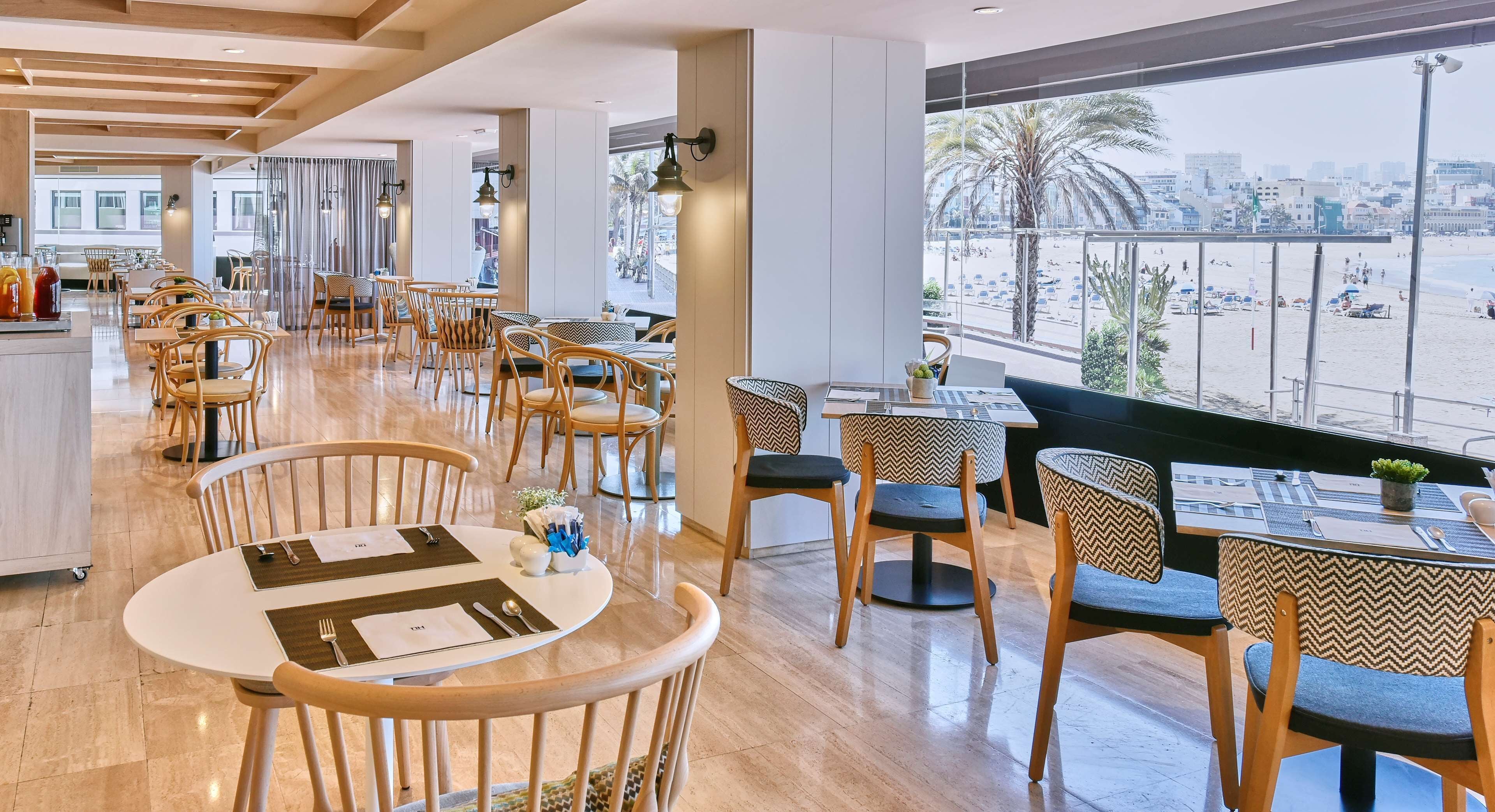 A modern restaurant with wooden chairs and tables, large windows offering a view of the beach and cityscape.