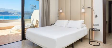 Habitación de hotel con una cama grande, dos lámparas de noche, una mesita pequeña y vista al mar.