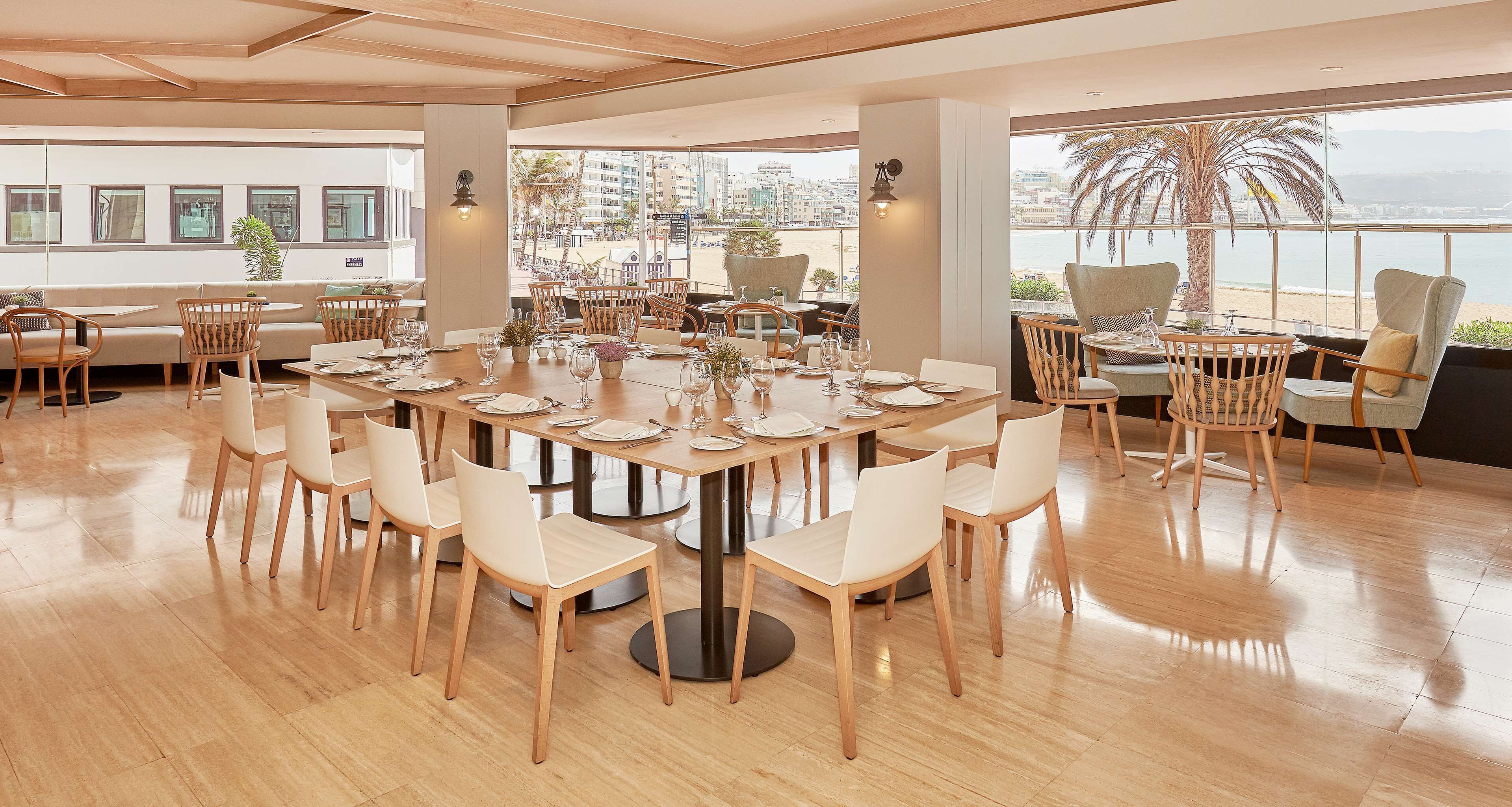 Un comedor amplio con una gran mesa redonda dispuesta para una comida, rodeada de sillas, y vistas a la ciudad y la playa a través de grandes ventanales.