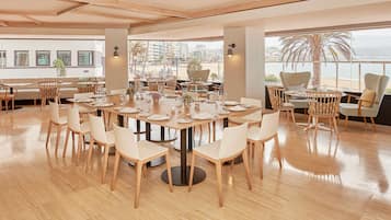 A spacious dining area with a large round table set for a meal, surrounded by chairs, with a view of the city and beach through large windows.