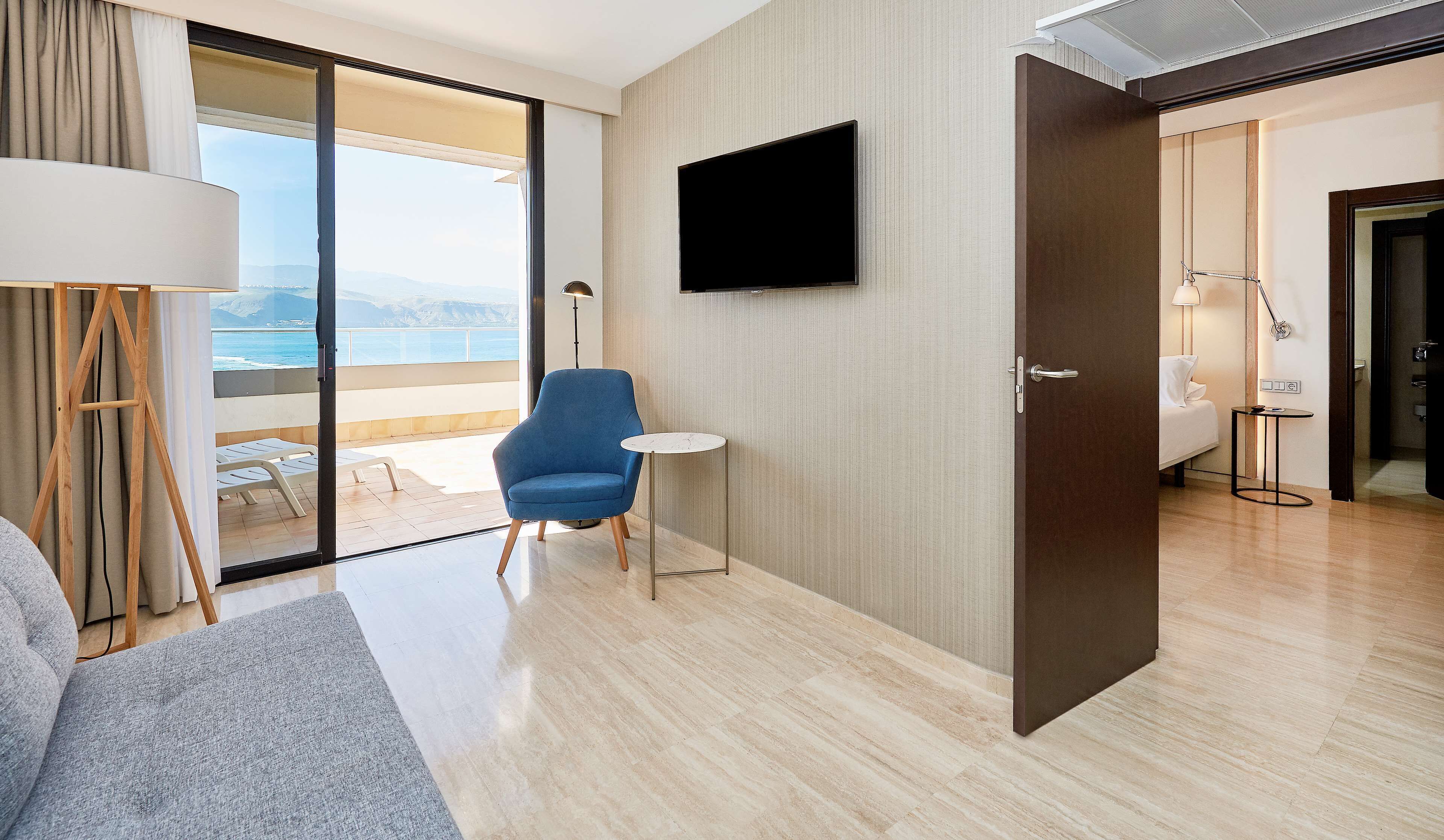 A modern hotel room with a large window, a flat-screen TV mounted on the wall, a blue armchair, a small round table, and a bed with a white headboard.