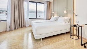 Habitación de hotel moderna con una cama grande, una mesita de noche con lámpara, vista al mar y suelo de madera.