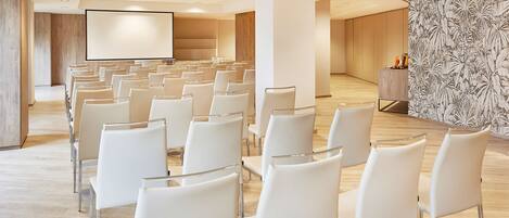 A conference room with rows of white chairs, a large screen, and a wooden panel wall.