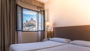 Una habitación de hotel con dos camas, una ventana que ofrece vistas a un castillo, una lámpara de noche y un teléfono en la mesita.
