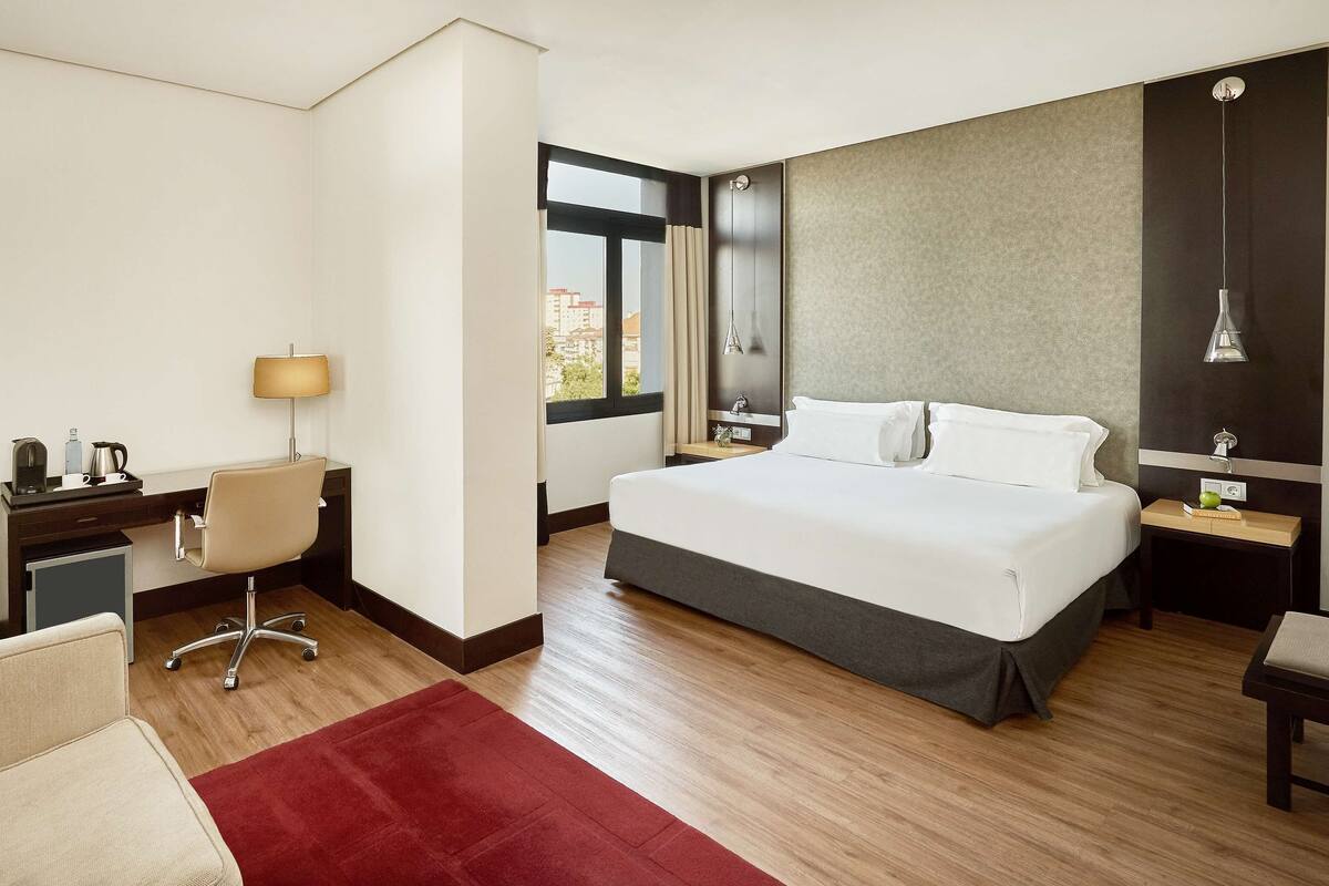 A hotel room with a bed, desk, chair, and a red rug.